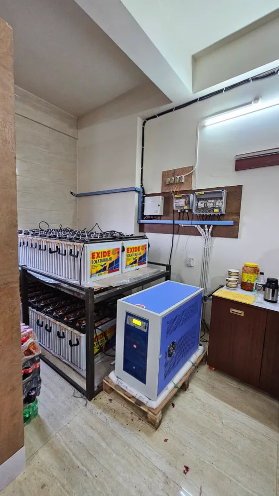 Two tiers of tubular batteries on a steel rack beside a blue inverter cabinet, with wall-mounted protection boxes and cable trunking.