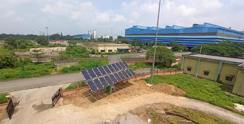 A ground-mounted array on a raised frame inside a fenced plot beside a road, large industrial buildings behind.