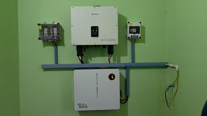 A wall-mounted inverter above a battery unit, with protection boxes on either side and cabling run through blue trunking on a green wall.