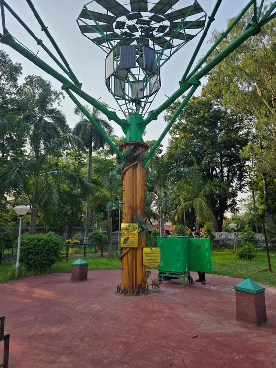 A green steel solar tree in a park, a canopy of modules raised on a sculpted trunk, with an equipment cabinet beside it.